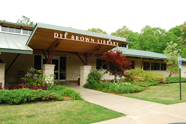 Covered entrance to single-story "Dee Brown Library" building with decorative plants in front yard