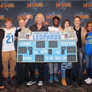 Five white men posing with mixed group of students holding up a scoreboard with "Def Leppard" logos in the background