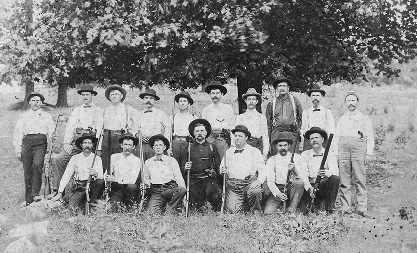 Group of white men with guns posing in field with trees in the background