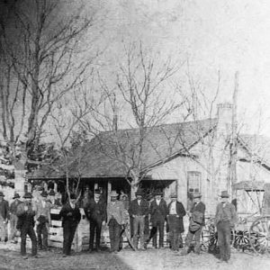 Group of white men in hats and suits standing outside single-story building hotel with carriages