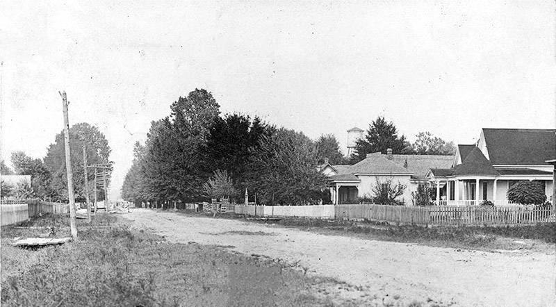 Houses with covered porches inside fence on dirt road with water tower in the background