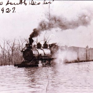Steam train on flooded tracks with smoke billowing from its smoke stack