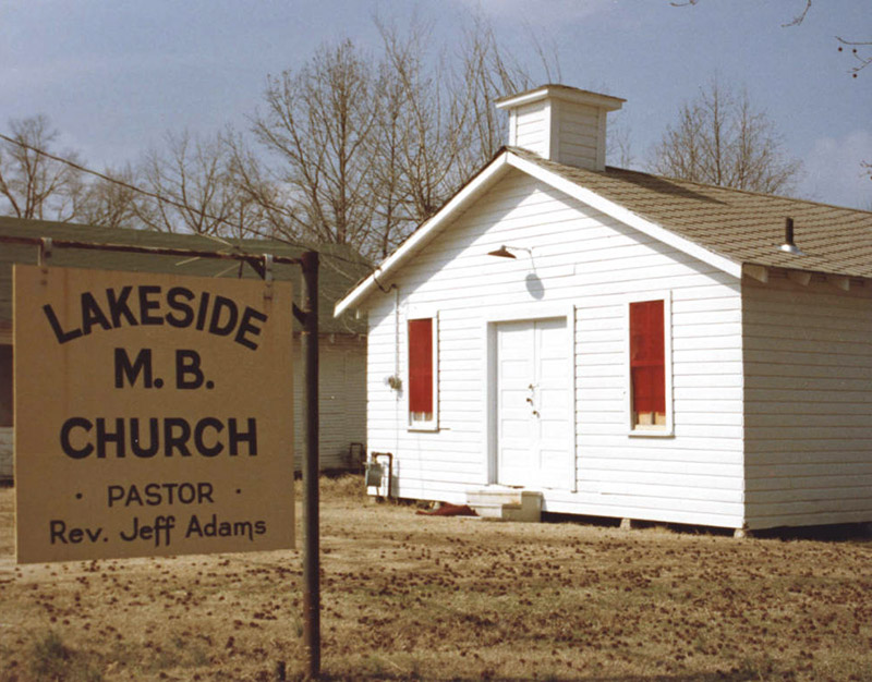 Single-story church building with cupola and wood siding with sign in the foreground