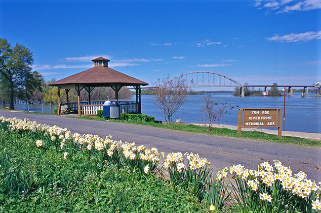 White flowers along walking path with pavilion and sign next to river with steel truss bridge over it in the distance
