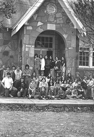 Group of white children and teenagers posing on steps of stone school house with arched entrance way