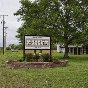 "Desha County Museum established 1979" sign in flower bed with log cabin and tree behind it