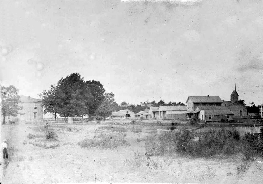 White man standing in field with town buildings and church on dirt roads in the background