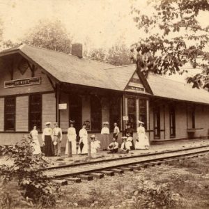 Crowd of white women and men at train depot building with tracks in the foreground