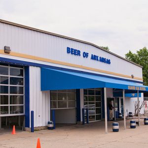 Metal "Beer of Arkansas" building with three garage bays and blue awning with parking lot