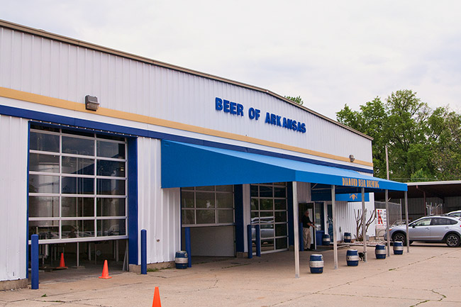 Metal "Beer of Arkansas" building with three garage bays and blue awning with parking lot