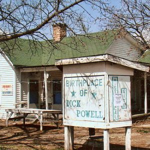 Single-story house with white siding tables and "Birthplace of Dick Powell" sign under trees