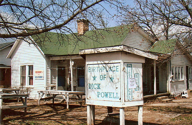 Single-story house with white siding tables and "Birthplace of Dick Powell" sign under trees