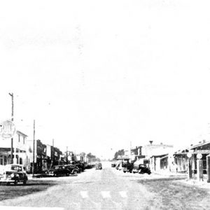 Esso gas station and storefronts on street with parked cars