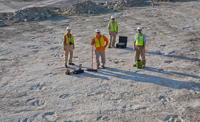 Four white men in hard hats and reflective vests with equipment in dirt field