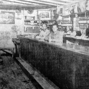 Two white men at a long wooden bar with bottles behind them