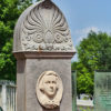 Bust of young white boy on engraved stone monument with trees and other monuments in the background