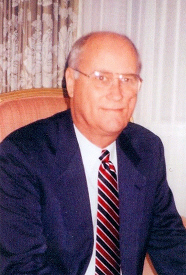 White man with glasses in suit and tie sitting and smiling in chair