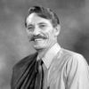 White man with mustache smiling in shirt and tie with suit jacket hanging over his right shoulder