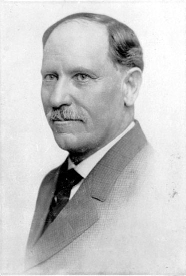 White man with mustache posing in suit and tie