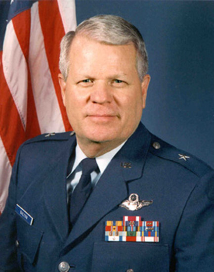 White man with gray hair in military uniform and flag behind him