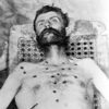 Corpse of white man with beard and multiple bullet wounds in bare chest being propped up in a chair