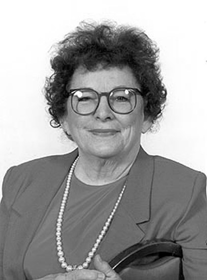 White woman smiling in glasses and suit with pearl necklace