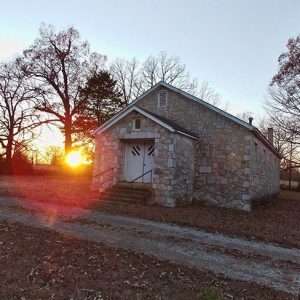 Single-story stone building with covered porch on dirt driveway with setting sun behind hit