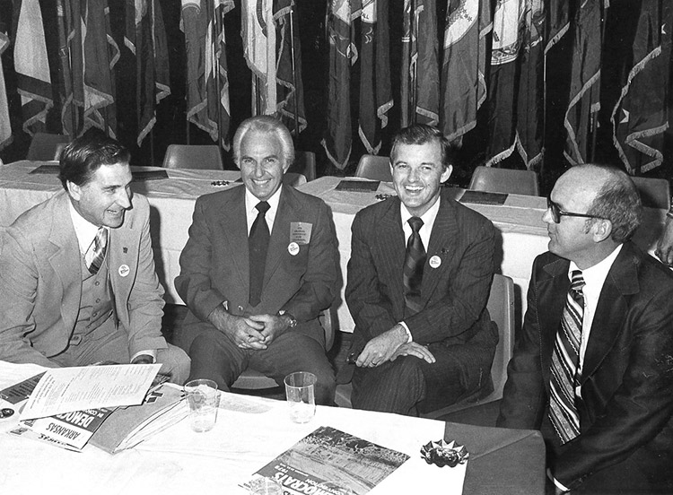 Four white men in suit and tie sitting and smiling at a table with row of flags behind them