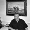 Old white man sitting at his desk with a framed painting of two Native American men on horseback hanging behind him