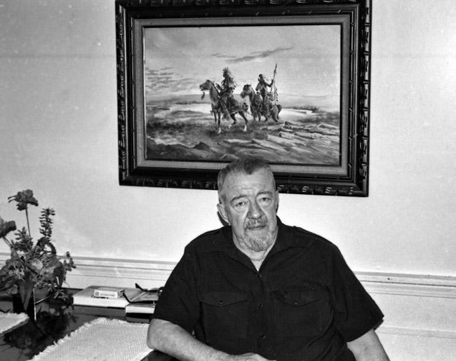 Old white man sitting at his desk with a framed painting of two Native American men on horseback hanging behind him