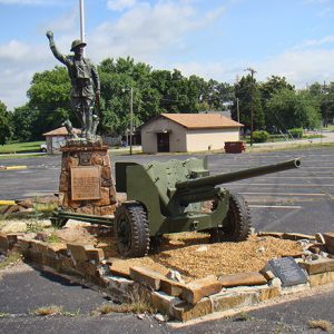 Soldier statue on brick pedestal with artillery piece and flag pole on parking lot with outbuildings in the background
