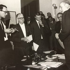 Group of white men in suits standing before white man in suit at desk in office