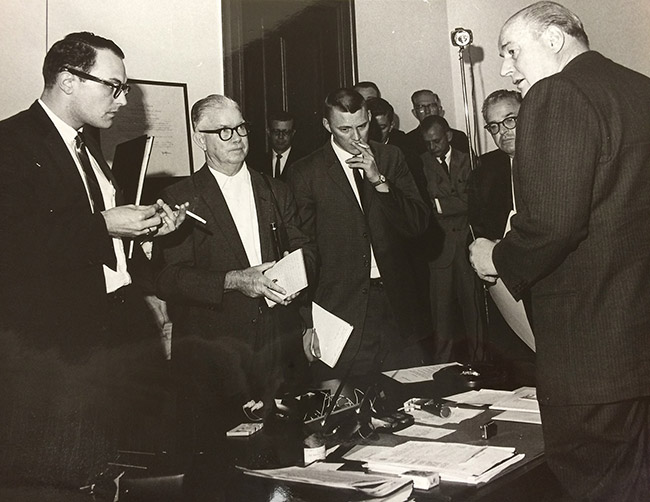 Group of white men in suits standing before white man in suit at desk in office