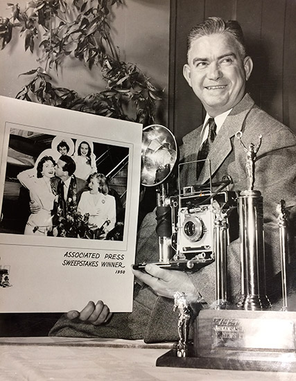 White man in suit smiling while holding camera and framed photograph with trophy in the foreground
