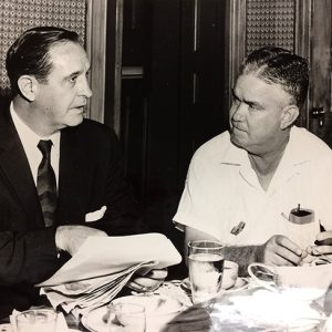 White man in suit and tie holding papers talking to white man in collared shirt at table