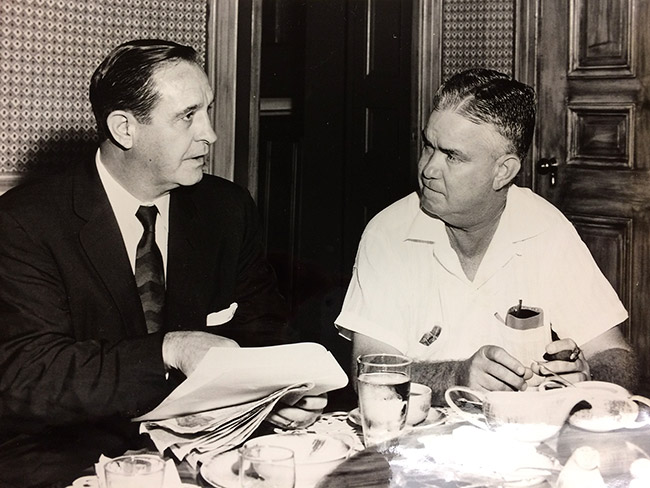 White man in suit and tie holding papers talking to white man in collared shirt at table