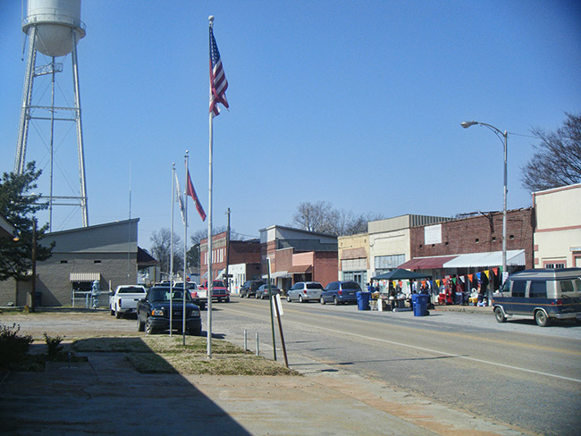 Street with brick storefronts flag poles parked cars and water tower