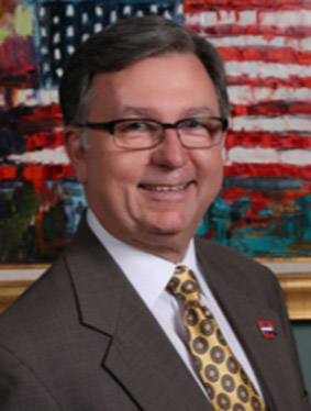 White man with glasses smiling in suit jacket and tie with flag behind him