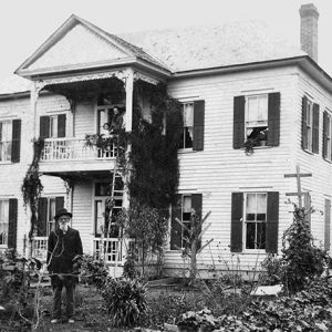 Older white man in hat and suit standing outside multistory house with covered porch and a balcony with people standing on it