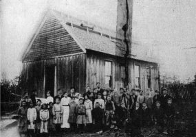 Group of white children and teachers posing for a photo outside single-story building with open gable roof