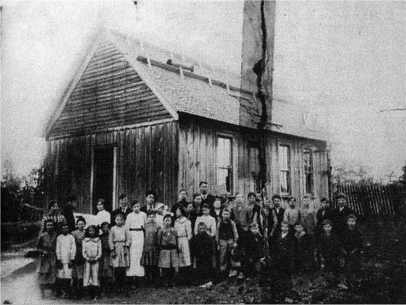 Group of white children and teachers posing for a photo outside single-story building with open gable roof