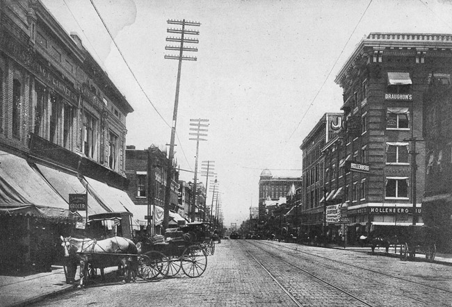 Street with trolley tracks lined with tall buildings electrical poles and lines and horse-drawn carriages