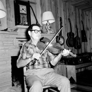 White man in glasses playing a violin/fiddle by the fireplace with guitars hanging on the wall in the background