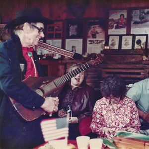 Old white man in hat and glasses with acoustic guitar performing for white patrons
