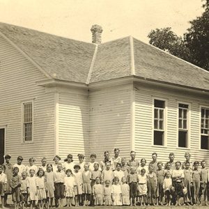 Group of white students and teachers posing for a group photo outside white wooden building
