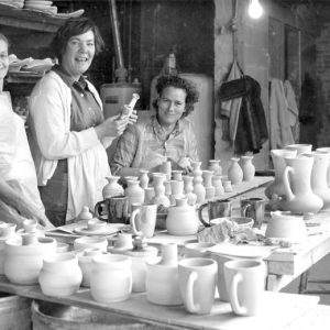Three white women in pottery factory with bowls in the background with small cups and jugs in front of them