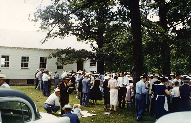 Crowd gathered under trees outside single-story building