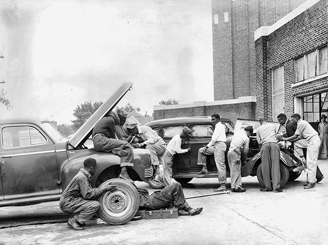 Group of young African American men working on cars outside multistory brick building