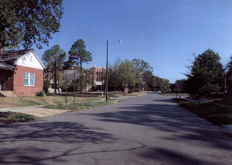 Brick house on street with multistory brick school building in the background behind trees