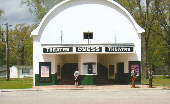 Green and white theater building with round roof on street with one person standing in front of the box office window